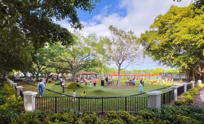 Warm sunny day, blue sky and clouds, people sit and play in the grass in the shade of large trees. In the foreground a fence enclosing the park.